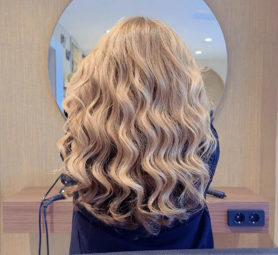 Long beige blonde hair with soft waves, photographed from behind in front of a round salon mirror
