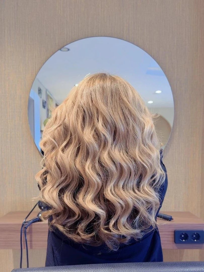 Lange beigeblonde Haare mit weichen Wellen, von hinten vor einem runden Spiegel im Salon fotografiert