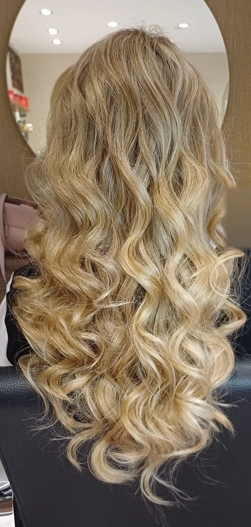 Lange goldblonde Haare mit großen Wellen und dunklerem Ansatz, von hinten vor einem runden Spiegel im Salon fotografiert