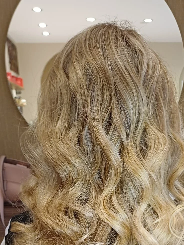 Lange goldblonde Haare mit großen Wellen und dunklerem Ansatz, von hinten vor einem runden Spiegel im Salon fotografiert