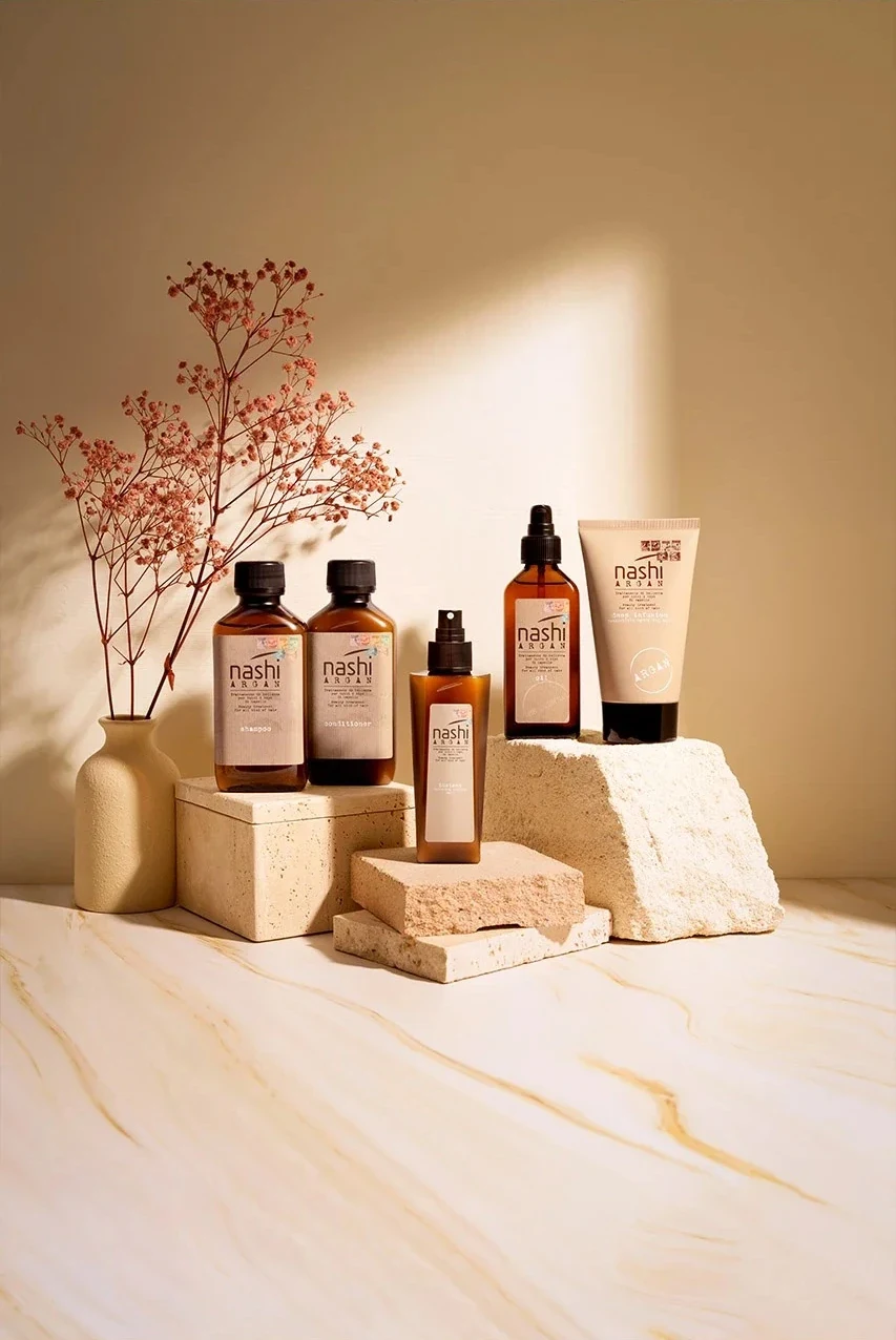 Pflegeprodukte von Nashi Argan: Flaschen und eine Tube auf hellem Stein vor einer beigen Wand